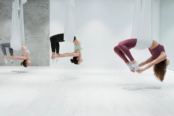 Vista lateral de tres mujeres jóvenes practicando yoga aéreo en el gimnasio - foto de stock