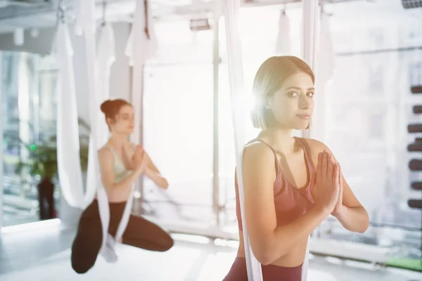 Mujer joven mirando la cámara mientras que practica la pose del loto en la hamaca aérea del yoga, fondo borroso - foto de stock