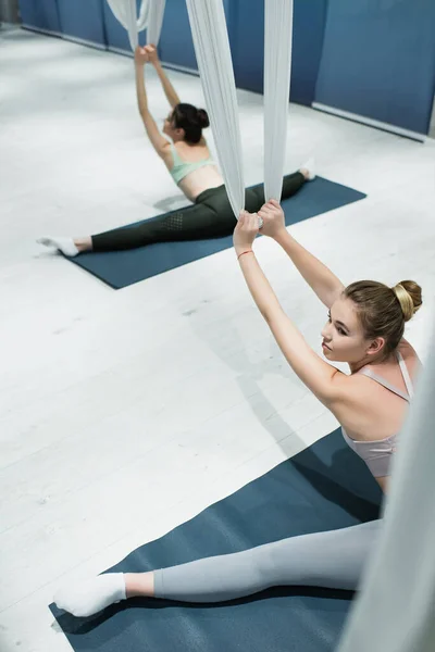 Vue grand angle de jeunes femmes s'étirant avec des sangles de yoga de mouche tout en étant assis sur des tapis de fitness — Photo de stock