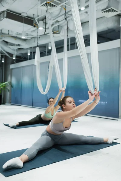Junge Sportlerinnen sitzen auf Fitnessmatten und dehnen sich mit Luft-Yoga-Hängematten — Stockfoto
