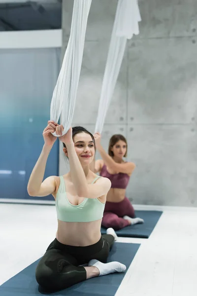 Deportista sonriente sentada en una colchoneta de fitness y sujetando correas de yoga con mosca durante el entrenamiento en grupo - foto de stock