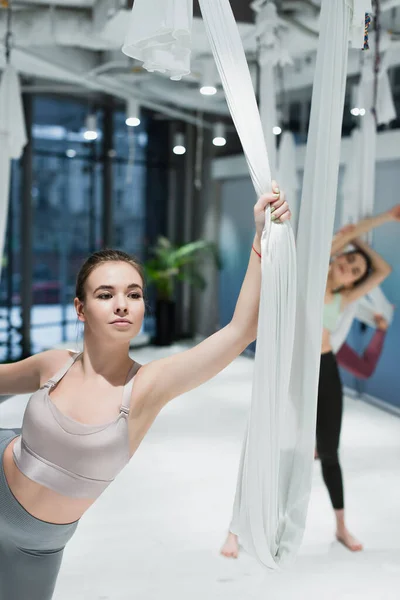 Mujer joven sosteniendo correas de yoga con mosca cerca de sportswomen sobre fondo borroso - foto de stock