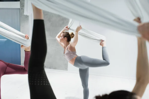 Side view of fit sportswoman training with fly yoga straps near women on blurred foreground — Stock Photo