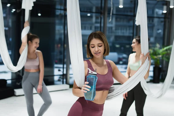 Mujer positiva mirando botella de deportes durante el entrenamiento de yoga con mosca - foto de stock