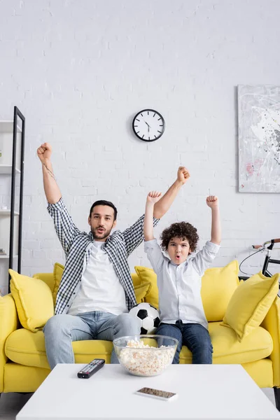 Emocionado niño árabe y padre mostrando gesto de éxito mientras ve el partido de fútbol en casa - foto de stock