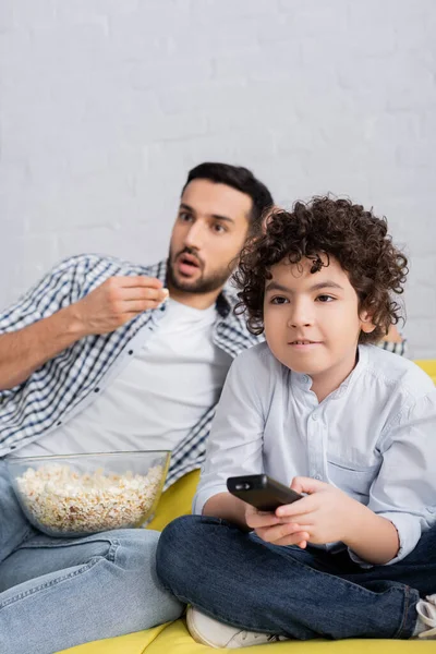 Concentrado arabian chico viendo tv cerca shocked padre comer palomitas de maíz en borrosa fondo - foto de stock