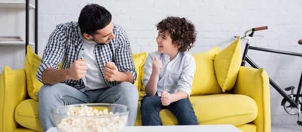 Happy arabian boy showing thumb up while talking to father on sofa at home, banner — Stock Photo