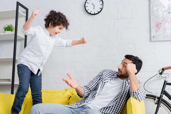 Surprised muslim man pointing at cheerful son jumping on sofa with clenched fists — Stock Photo
