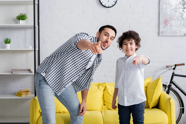 Cheerful arabian man and son sticking tongues and pointing with fingers while looking at camera — Stock Photo