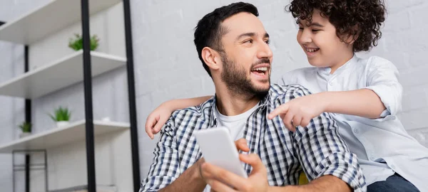 Веселий мусульманський хлопчик вказує на смартфон в руках усміхненого батька, банер — стокове фото