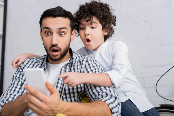 Astonished muslim boy pointing at smartphone near shocked father on blurred foreground — Stock Photo