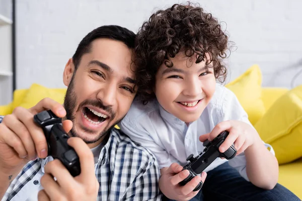 KYIV, UKRAINE - JANUARY 15, 2021: astonished muslim man playing video game with cheerful son at home — Stock Photo