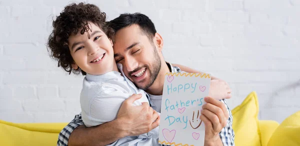 Hombre árabe complacido con feliz tarjeta del día del padre abrazando hijo alegre, bandera - foto de stock