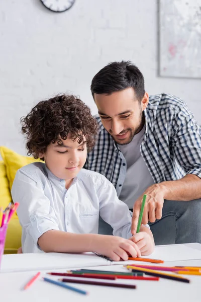 Homme arabe souriant pointant du doigt près du fils dessin au crayon, avant-plan flou — Photo de stock