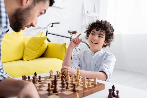 Menino mulsim alegre apontando com o dedo enquanto joga xadrez com o pai em primeiro plano borrado — Fotografia de Stock