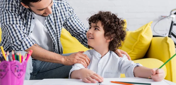 Niño musulmán feliz mirando a padre mientras que sostiene el lápiz cerca del cuaderno de bocetos, primer plano borroso, bandera — Stock Photo