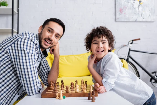 Cheerful arabian father and son looking at camera near chess on table — Stock Photo