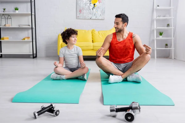 Sourire arabe père et fils assis dans la pose de yoga près des haltères à la maison — Photo de stock