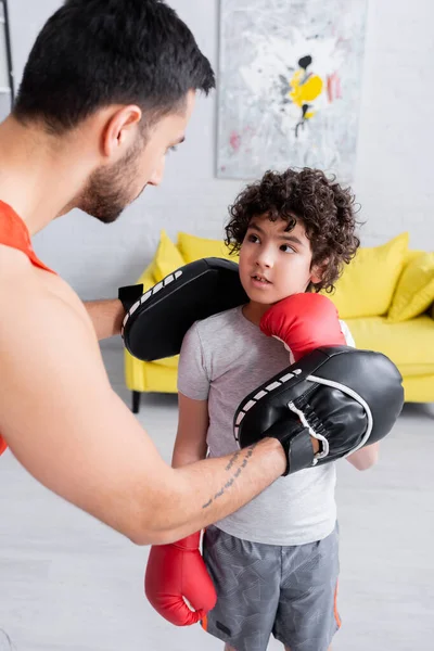 Musulmán padre en guantes de ponche enseñando hijo en guantes de boxeo en casa - foto de stock