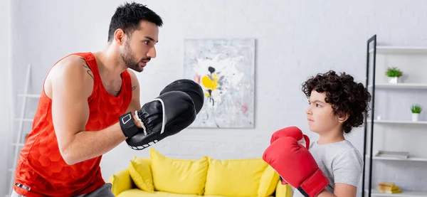 Hijo musulmán en guantes de boxeo de pie cerca del padre durante el trabajo en casa, pancarta - foto de stock