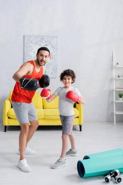 Sonriente padre e hijo árabe en guantes de boxeo sonriendo a la cámara cerca del equipo deportivo en casa - foto de stock