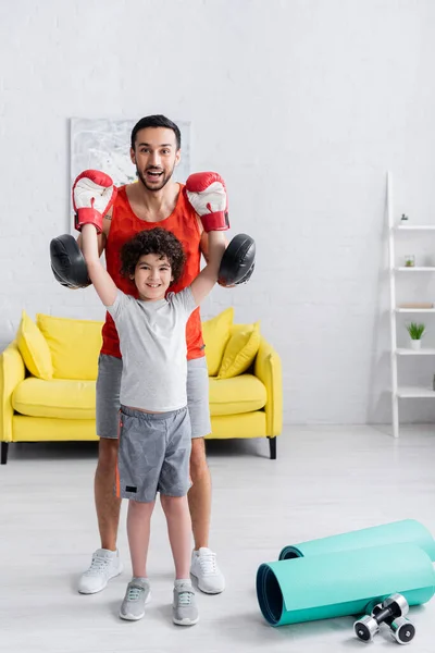 Árabe padre e hijo en guantes de boxeo sonriendo a la cámara en casa - foto de stock