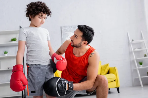 Hombre musulmán en guantes de ponche de pie cerca de hijo en guantes de boxeo en la sala de estar - foto de stock