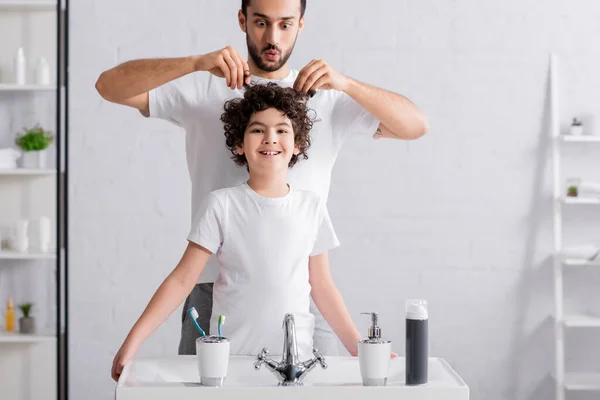 Cheerful muslim boy standing near father touching hair in bathroom — Stock Photo