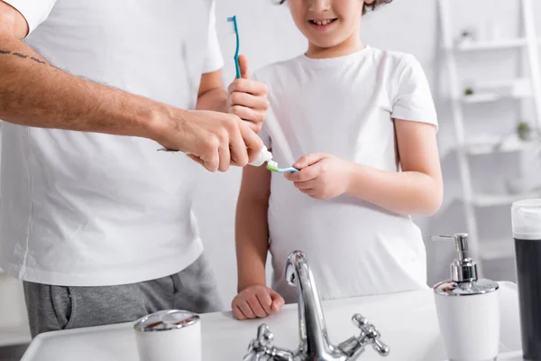 Cropped view of father applying toothpaste on toothbrush near smiling son in bathroom — Stock Photo