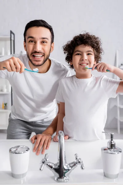 Arabian man holding toothbrush near son and sink on blurred foreground — Stock Photo
