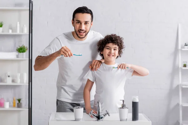 Smiling muslim man looking at camera near son with toothbrush at bathroom — Stock Photo