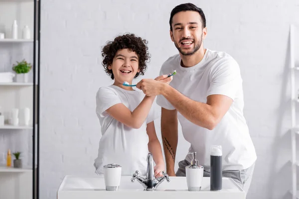 Heureux fils et père musulman tenant brosses à dents et souriant à la caméra dans la salle de bain — Photo de stock