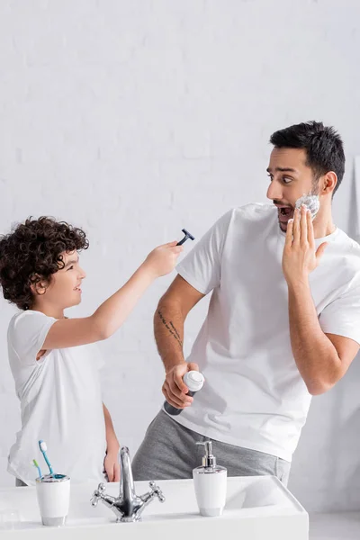 Smiling arabian son holding razor near scared father with shaving foam — Stock Photo