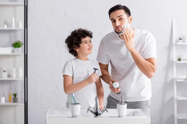 Smiling arabian boy holding razor near father with shaving foam in bathroom — Stock Photo