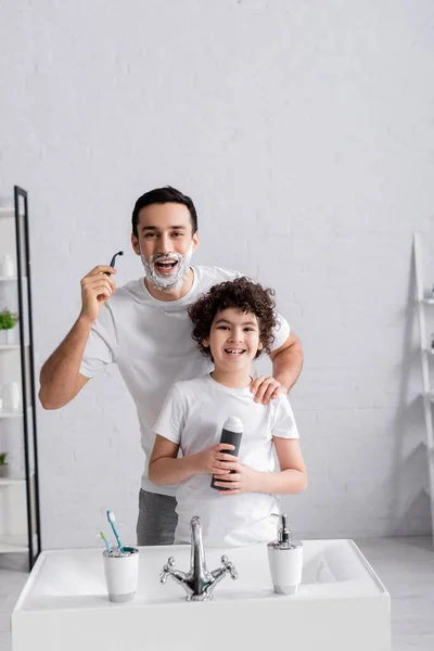 Cheerful arabian man holding razor near son with shaving foam — Stock Photo