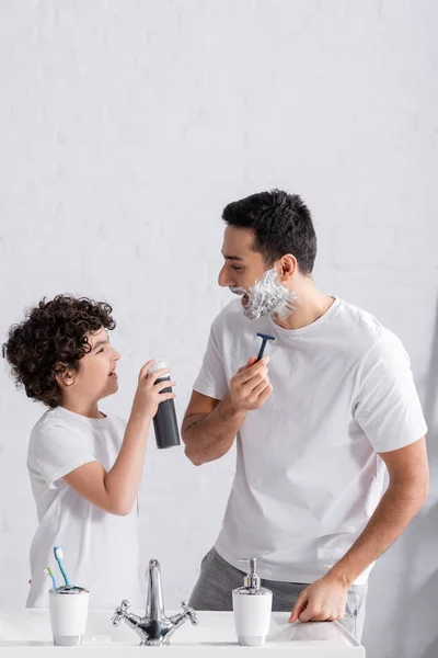 Smiling muslim boy holding shaving foam near father with razor and sink — Stock Photo