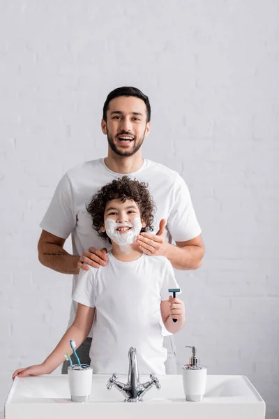 Smiling arabian father applying shaving foam on face of son with razor in bathroom — Stock Photo