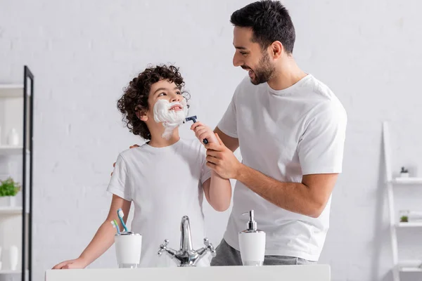 Sonriente hombre musulmán ayudando a su hijo en espuma de afeitar con afeitadora - foto de stock