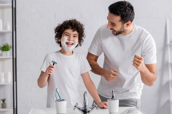Hombre alegre de pie cerca de hijo con espuma de afeitar y maquinilla de afeitar en el baño - foto de stock