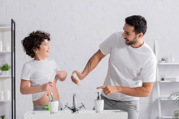 Smiling arabian man holding deodorant near son and sink in bathroom — Stock Photo