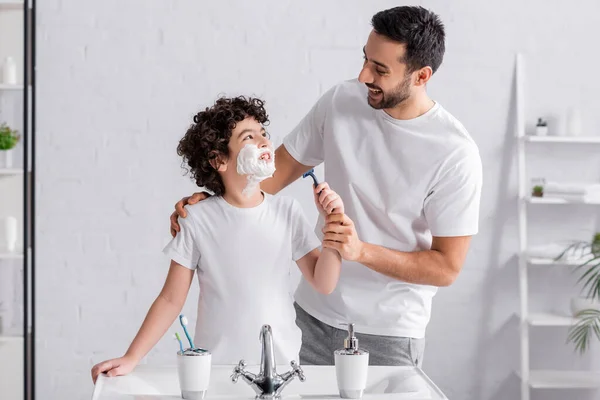 Muslim boy with shaving foam on face holding razor and looking at father — Stock Photo