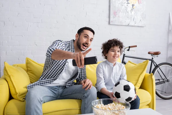 Araber mit Fernbedienung zeigt mit Finger auf Popcorn und Sohn mit Fußball zu Hause — Stockfoto