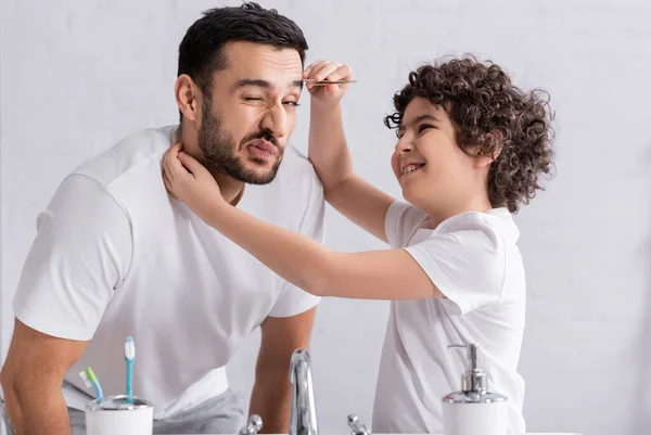 Sonriente chico musulmán sacando la ceja del padre en el baño - foto de stock