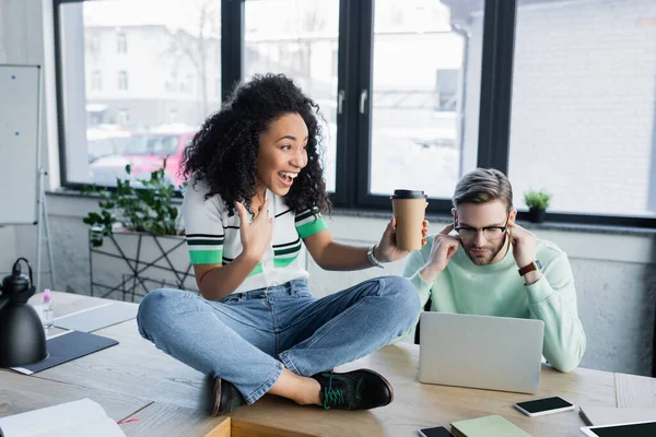 Positiva mujer de negocios afroamericana con café para ir sentado en la mesa cerca de colegas cubriendo las orejas — Stock Photo