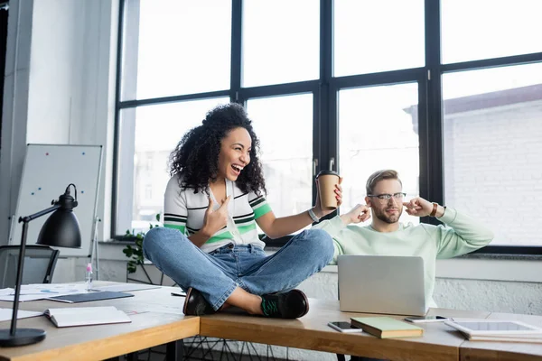 Empresario cubriendo oídos cerca de alegre colega afroamericano con café para ir a la mesa de la oficina — Stock Photo
