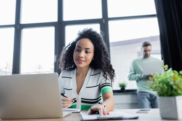 Smiling african american businesswoman working with clipboard and laptop on blurred foreground — Stock Photo