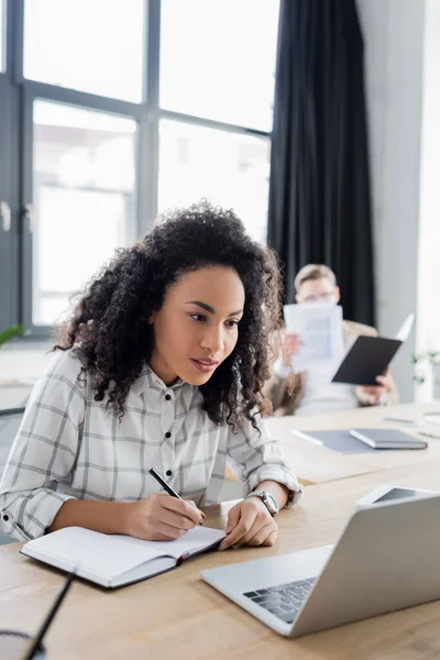 African american businesswoman writing on notebook near laptop on blurred foreground in office — Stock Photo