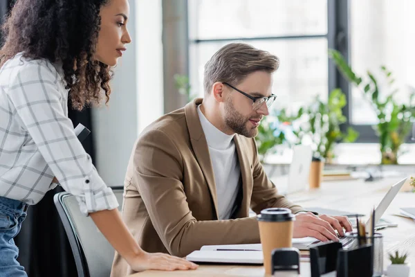 Multiethnische Geschäftsleute mit Laptop in der Nähe von Coffee to go und Schreibwaren im verschwommenen Vordergrund — Stockfoto