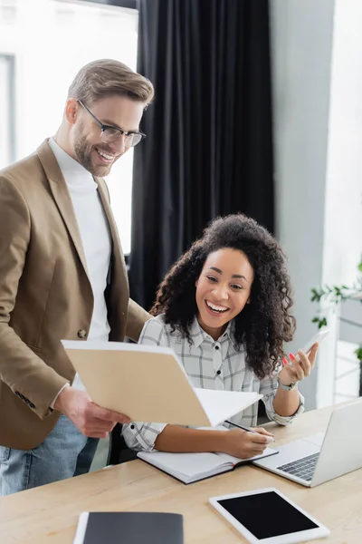 Hombre de negocios sonriente sosteniendo una carpeta de papel cerca de un colega afroamericano con teléfono inteligente y portátil - foto de stock