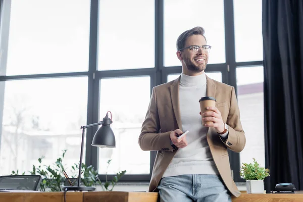Щасливий бізнесмен, дивлячись далеко, тримаючи смартфон і каву, щоб піти в офіс — стокове фото